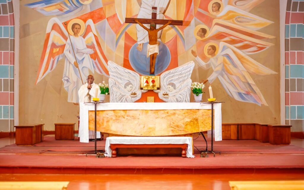 A priest stands at an ornately decorated church altar with religious art and a crucifix in the background.