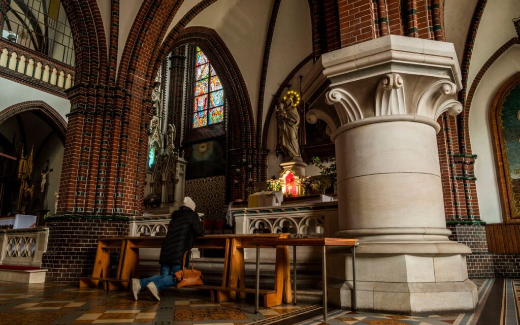 A person kneeling in prayer inside a historic brick cathedral with stained glass windows.